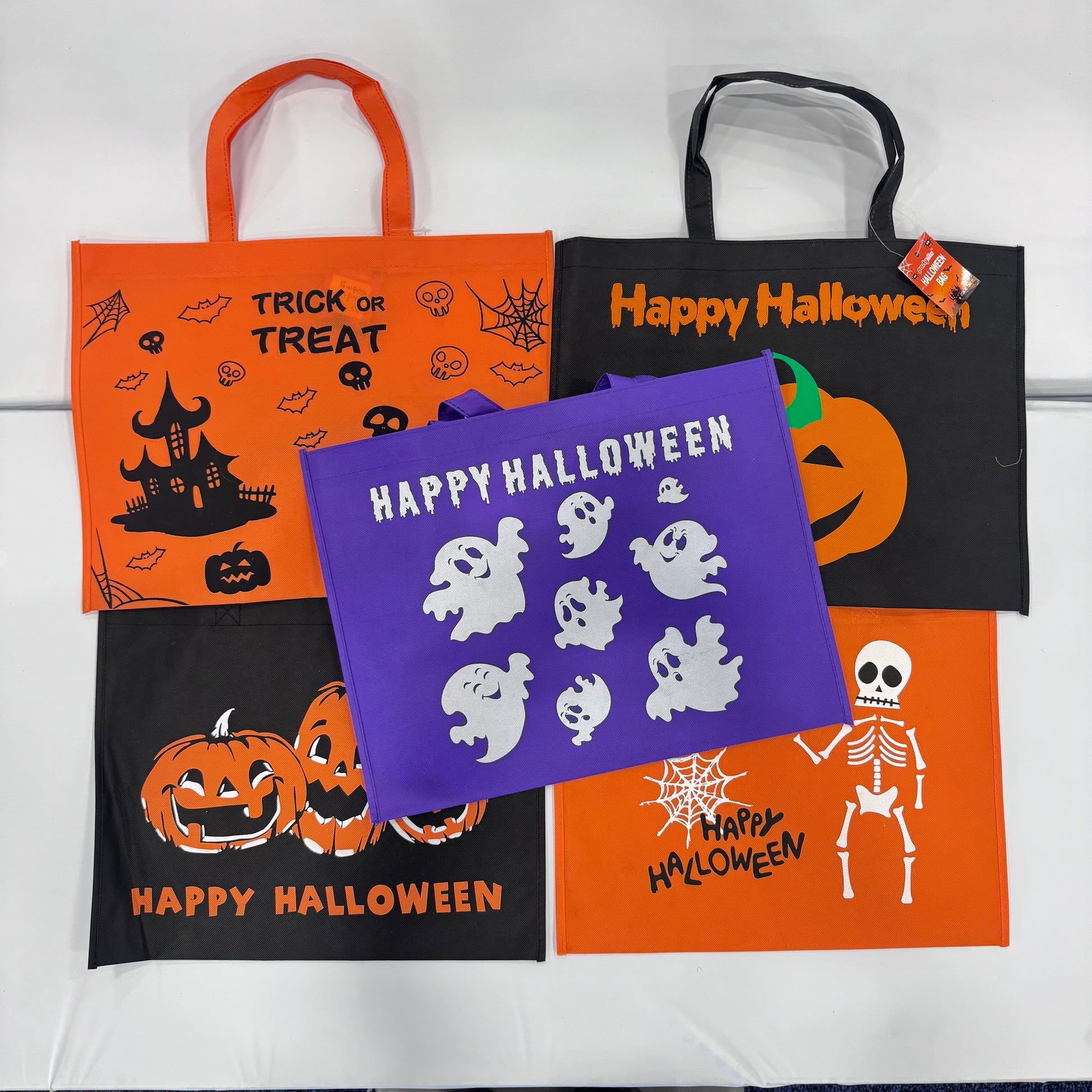Four Halloween-themed fabric tote bags arranged on a white background: orange with black handles and spooky graphics, black with orange handles and 'Happy Halloween' text, purple with white ghost cutouts and 'Happy Halloween' text, and orange with black handles, jack-o'-lantern graphics, and 'Happy Halloween' text.