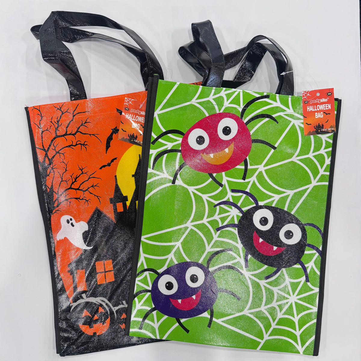 Two Halloween-themed plastic trick or treat bags with black handles, shown on a white background. One bag has an orange haunted house and ghost design, and the other has a green spider web design with three colorful cartoon spiders.
