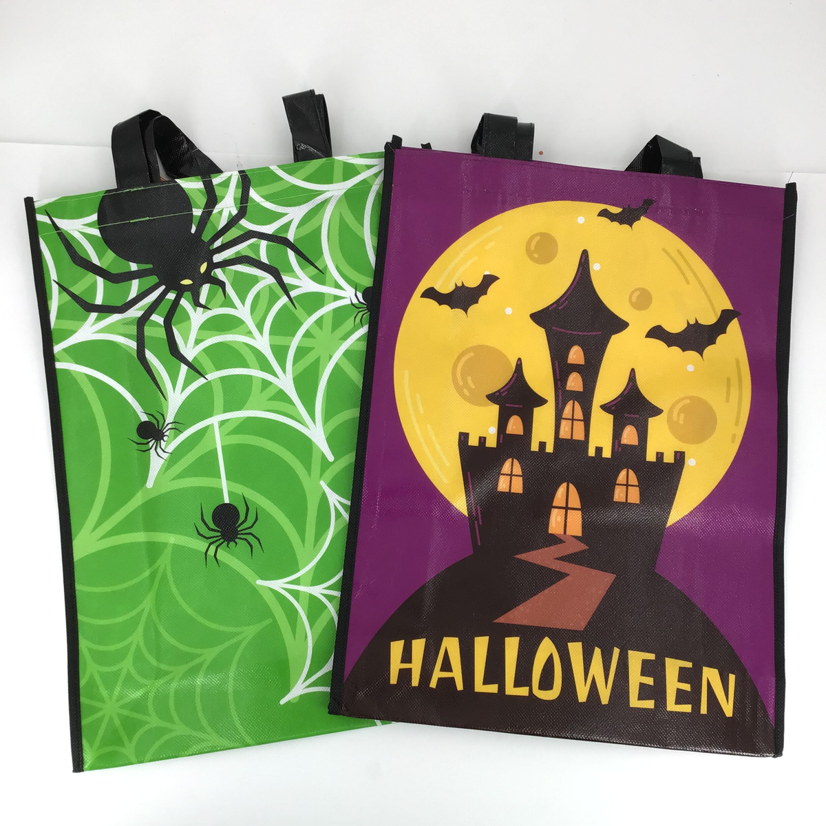 Two reusable Halloween shopping bags shown on a white background. One bag is bright green with black spider and white cobweb graphics, and the other is purple with a black haunted castle, yellow full moon, bats, and the word HALLOWEEN in yellow text.