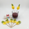 Pineapple-themed cocktail stirrers with a glass of red drink on a white background
